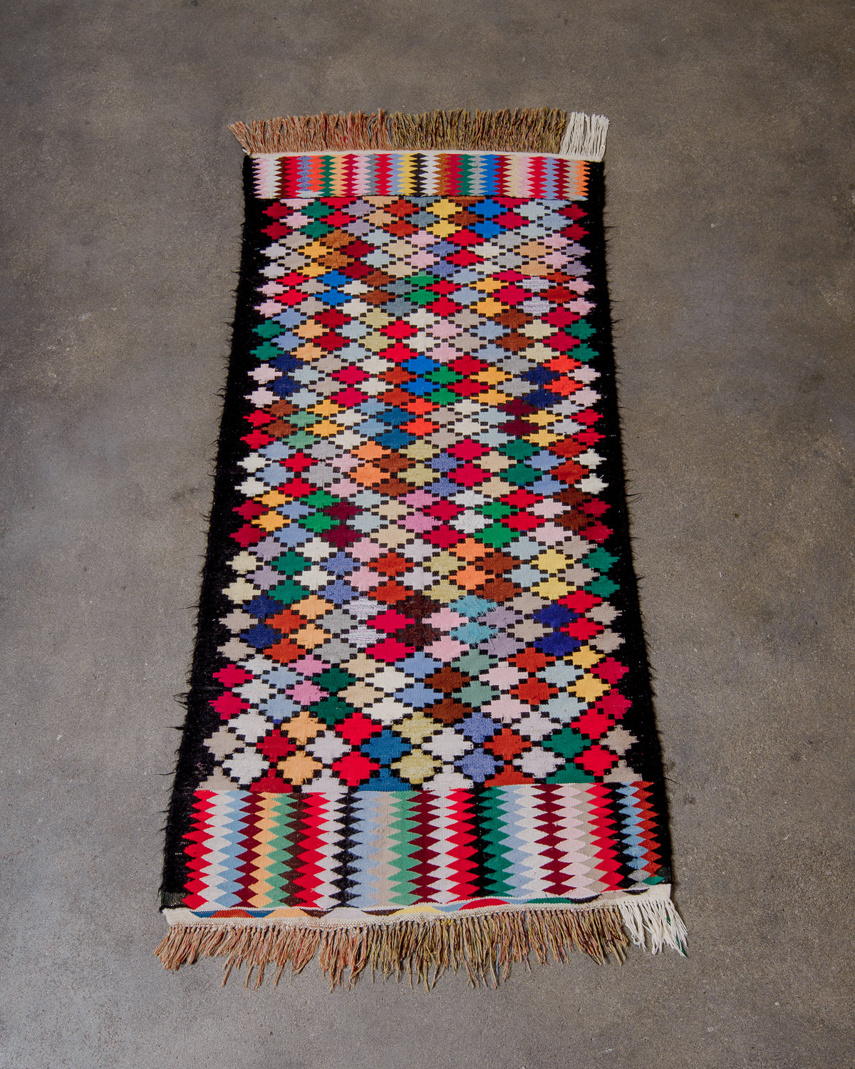 Berberlin Vintage Kelim runner rug with a brightly coloured design, black borders, and cotton tassels at each end, laying on concrete floor.